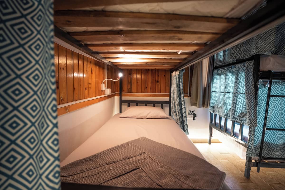 Una habitación pequeña y acogedora con paredes de madera y una cama individual sobre otra. Hay una mesita de noche con una lámpara y una ventana con cortinas estampadas.