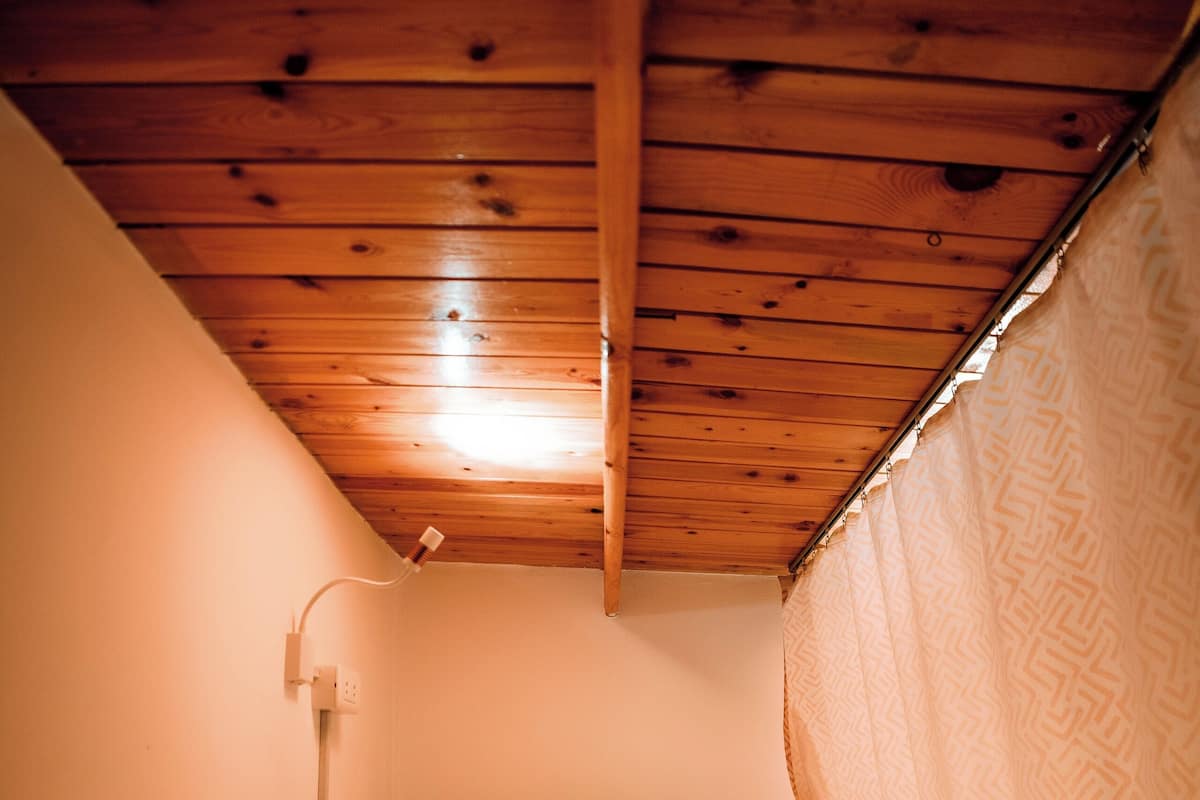 Una habitación pequeña con una cama individual, techo de madera, lámpara de noche y cortina estampada.
