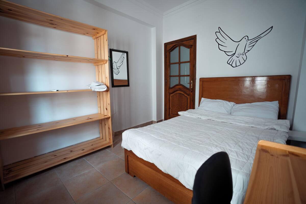 Un dormitorio con una cama de madera, una estantería de madera, un cuadro enmarco y un adhesivo mural con forma de pájaro.