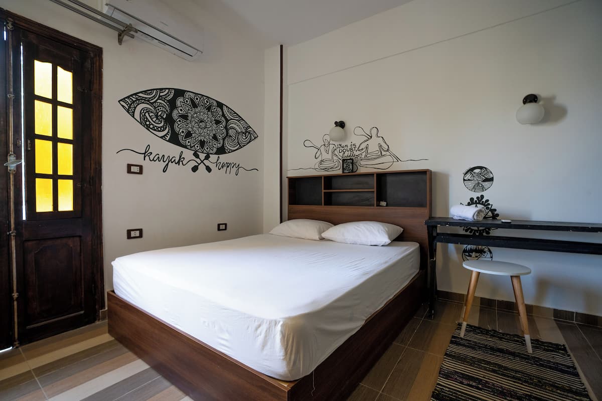 Un dormitorio con una cama de madera, un escritorio con silla, una pared decorada con arte y una puerta con ventana.