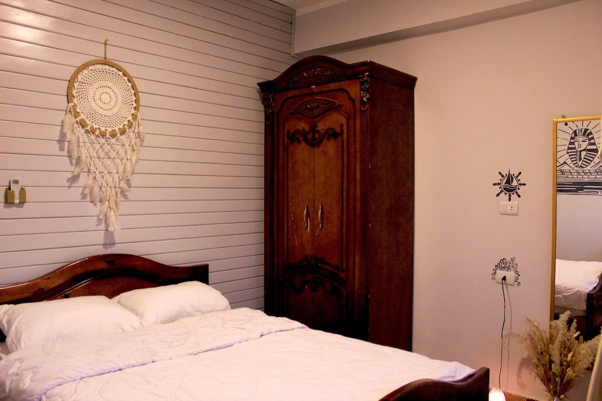 Un dormitorio con un armario de madera, una cama con ropa de cama blanca, un atrapasueños en la pared y un enchufe de pared.