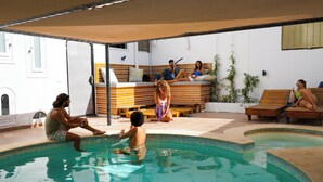 Un área de piscina con bordeado de azulejos, tumbonas de madera y una zona de estar cubierta con cojines.