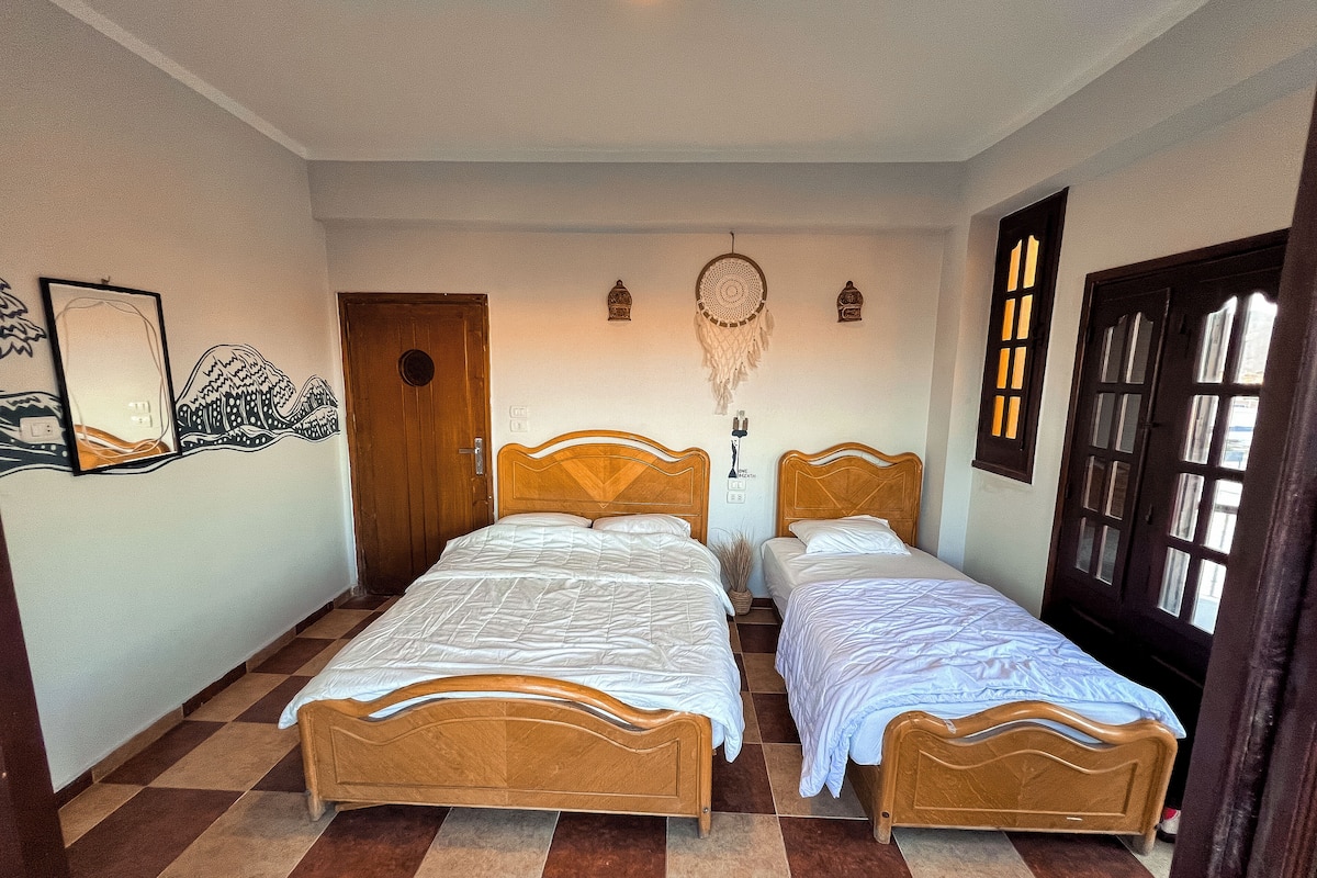 Una habitación con dos camas de madera, un suelo a cuadros, una puerta de madera y una pared decorada con un atrapasueños y un cuadro enmarcado.