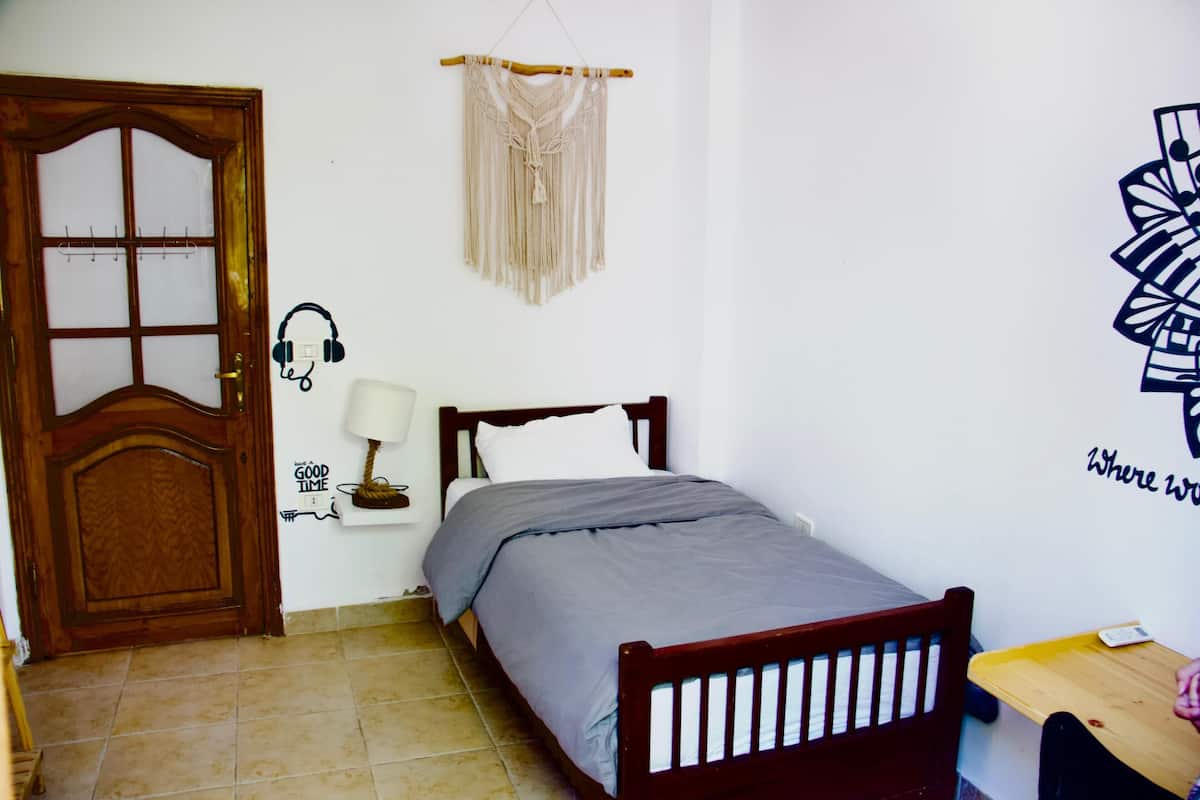 Un dormitorio con cama de madera, una mesita de noche pequeña, un adorno de macramé en la pared, una puerta con perchero para toallas y una pared con texto parcial y un gráfico.