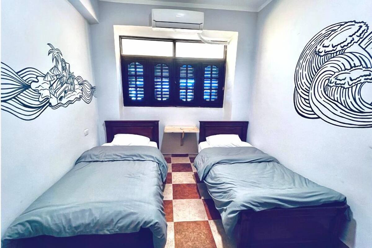 Una habitación pequeña con dos camas individuales, una ventana con vidrio azulado y decoración en la pared.