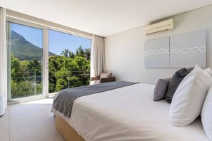 A bedroom with a large bed, a view of greenery and mountains, an air conditioning unit, and a balcony with a railing.