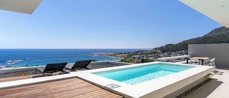 A rooftop pool with a view of the sea, a wooden deck, and a table with chairs.