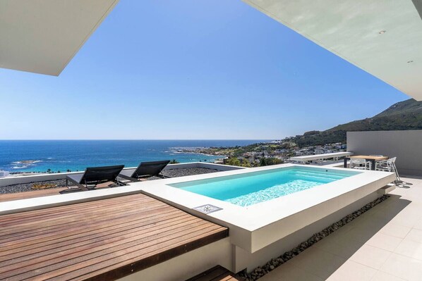 A rooftop pool with a view of the sea, a wooden deck, and a table with chairs.