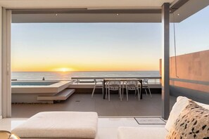 Een balkon met uitzicht op de ondergaande zon boven de oceaan, een zwembad en een zitgedeelte met stoelen.