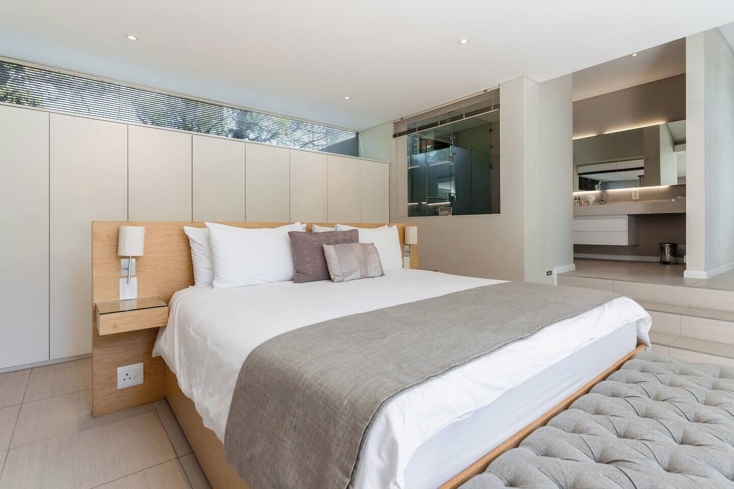 A modern bedroom with a large bed, bedside tables, a built-in wardrobe, and a bathroom visible through a glass door.