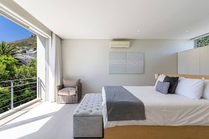 Un dormitorio moderno con una cama grande, un banco, una silla y vistas a zonas verdes y edificios.