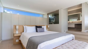 A modern bedroom with a large bed, a bedside table, a nightstand, a built-in wardrobe, and a view of the sky through a window. - Aquatic Penthouse - Coastal Luxury & Rooftop Views (Cape Town)