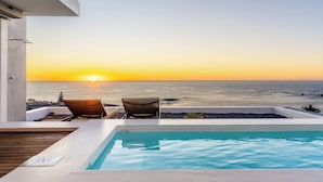 A poolside view with a sunset over the ocean, two chairs, and a modern building structure. - Aquatic Penthouse - Coastal Luxury & Rooftop Views (Cape Town)