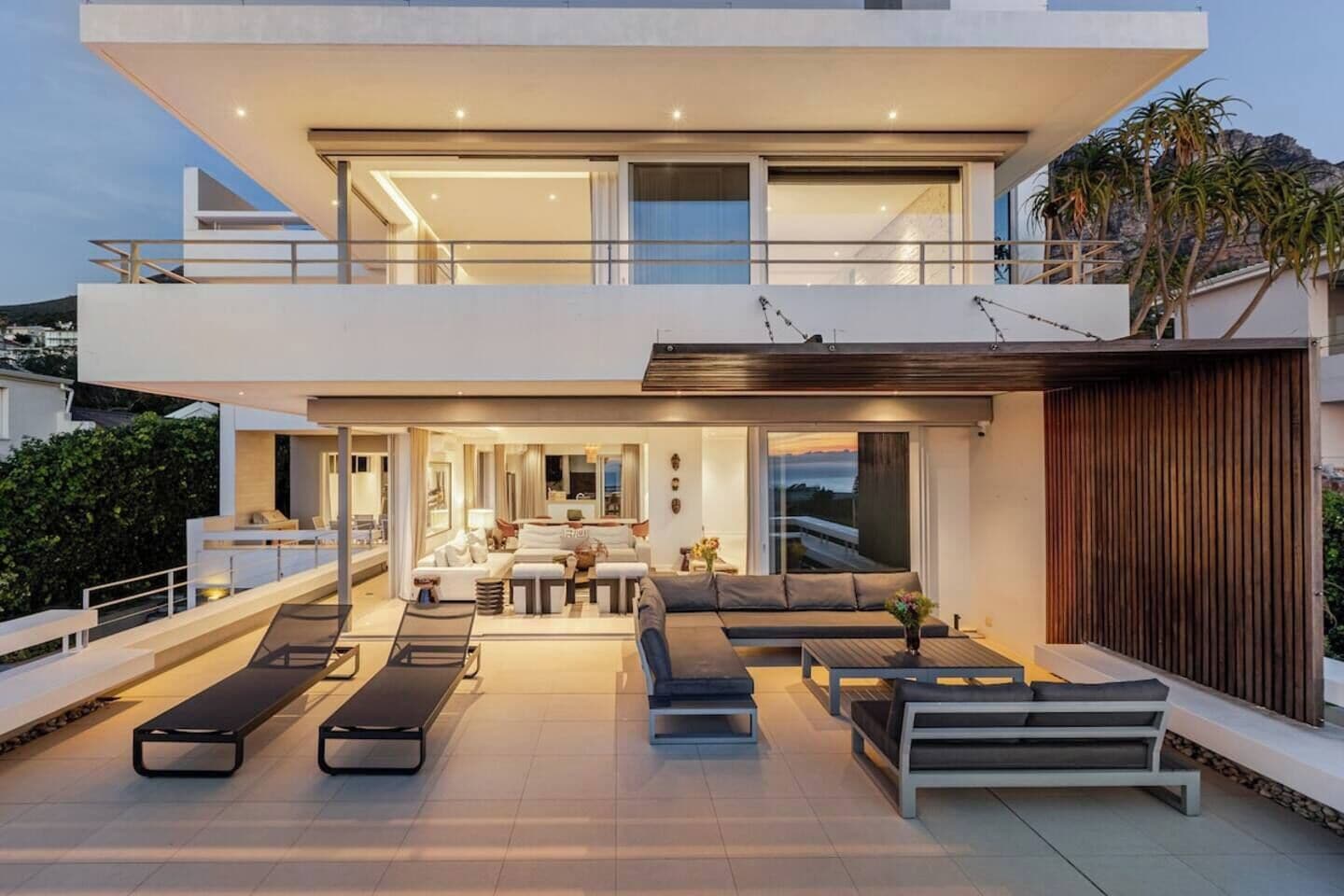 A modern two-story house with a balcony, outdoor seating, and a view of the landscape.