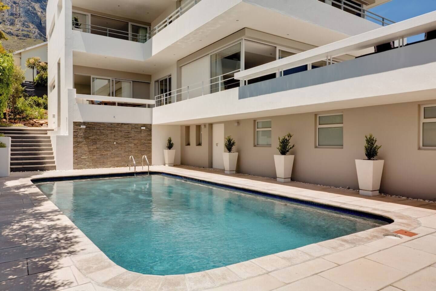A modern two-story building with a swimming pool, outdoor seating, and a stone wall.