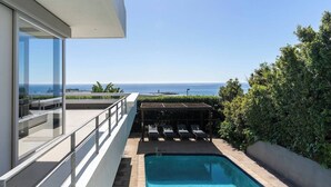 Piscina no terraço com vista para o oceano, área de estar coberta e guarda-corpo de vidro.