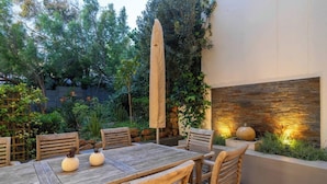 Um pátio externo com mesa e cadeiras de madeira, cercado por vegetação e um muro decorativo.