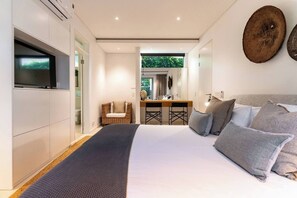 A modern bedroom with a large bed, a television, a dining area, and a view of greenery outside.
