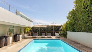 Ein Swimmingpool mit Liegestühlen und Sonnenschirmen, umgeben von einer Hecke und einem Gebäude.