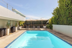 A swimming pool with lounge chairs and umbrellas, surrounded by a hedge and a building.