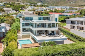 A modern two-story house with a pool, surrounded by lush greenery and other residential buildings.