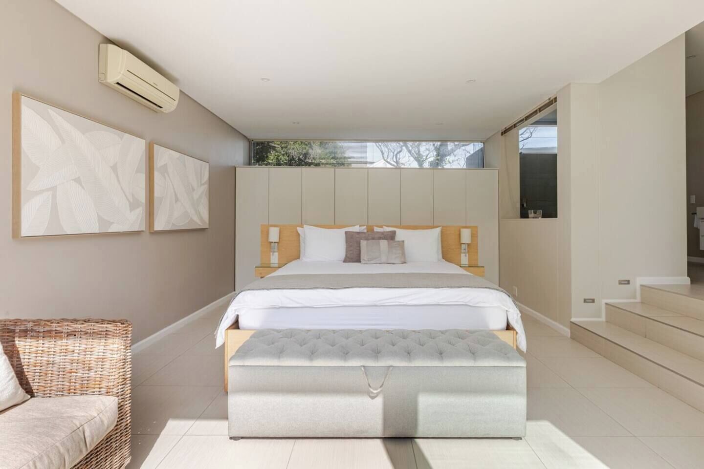 A modern bedroom with a large bed, a bedside table, a wall-mounted air conditioner, and a view of the outdoors.