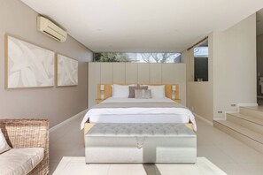 A modern bedroom with a large bed, a bedside table, a wall-mounted air conditioner, and a view of the outdoors.