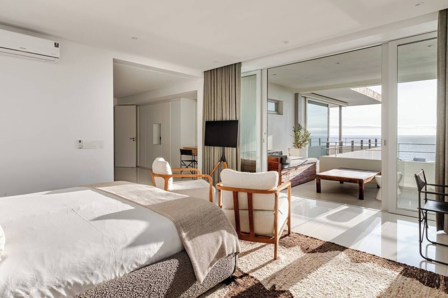 Une chambre d’hôtel moderne dotée d’un grand lit, d’un coin salon avec des fauteuils, d’une télévision à écran plat et d’un balcon offrant une vue sur la mer.