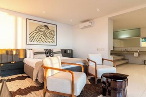 Ein modernes Hotelzimmer mit einem Bett, zwei Sesseln, einem Beistelltisch, einem Teppich, einem Gemälde und einer Klimaanlage, mit Blick ins Badezimmer.
