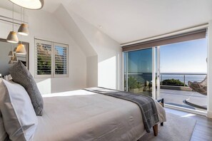 A bedroom with a large bed, a view of the sea through a sliding glass door, and a balcony with a chair.
