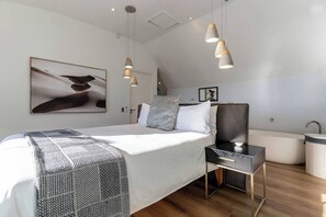 A modern bedroom with a large bed, bedside tables, a nightstand, a wall-mounted artwork, a hanging light fixture, and a built-in bathtub.