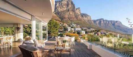 A modern outdoor deck with wicker furniture, a glass balustrade, and a view of a mountain and town.