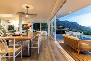 A modern interior with a wooden dining table, white chairs, and a wicker sofa on a deck with a mountain view.