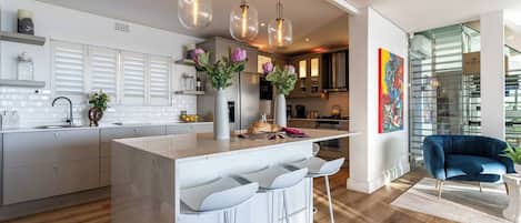 Una cocina moderna con una isla central, taburetes de bar, iluminación colgante y vistas al área de comedor.
