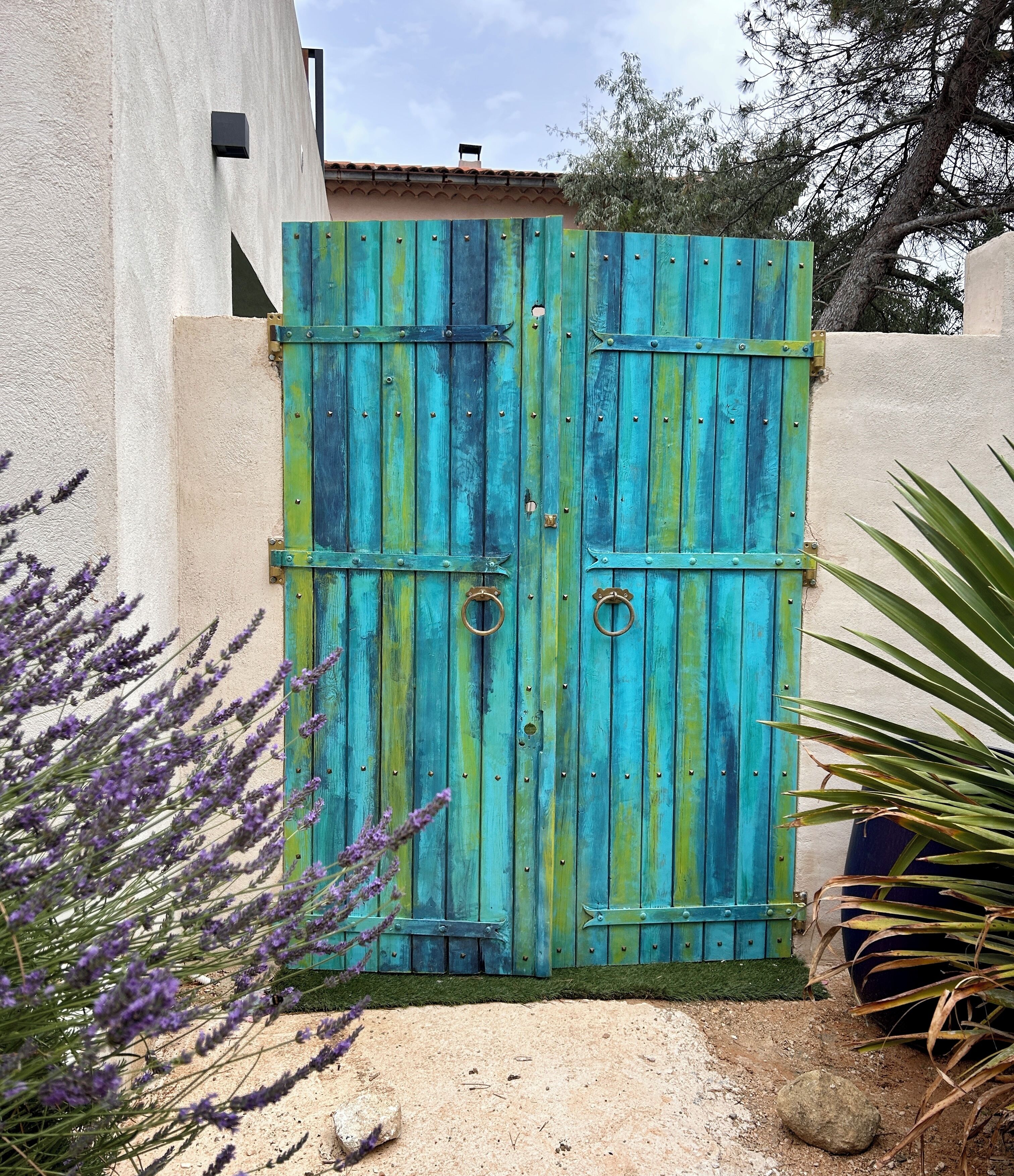 A rustic wooden gate with metal hinges and a circular handle, set against a beige wall with a textured surface.