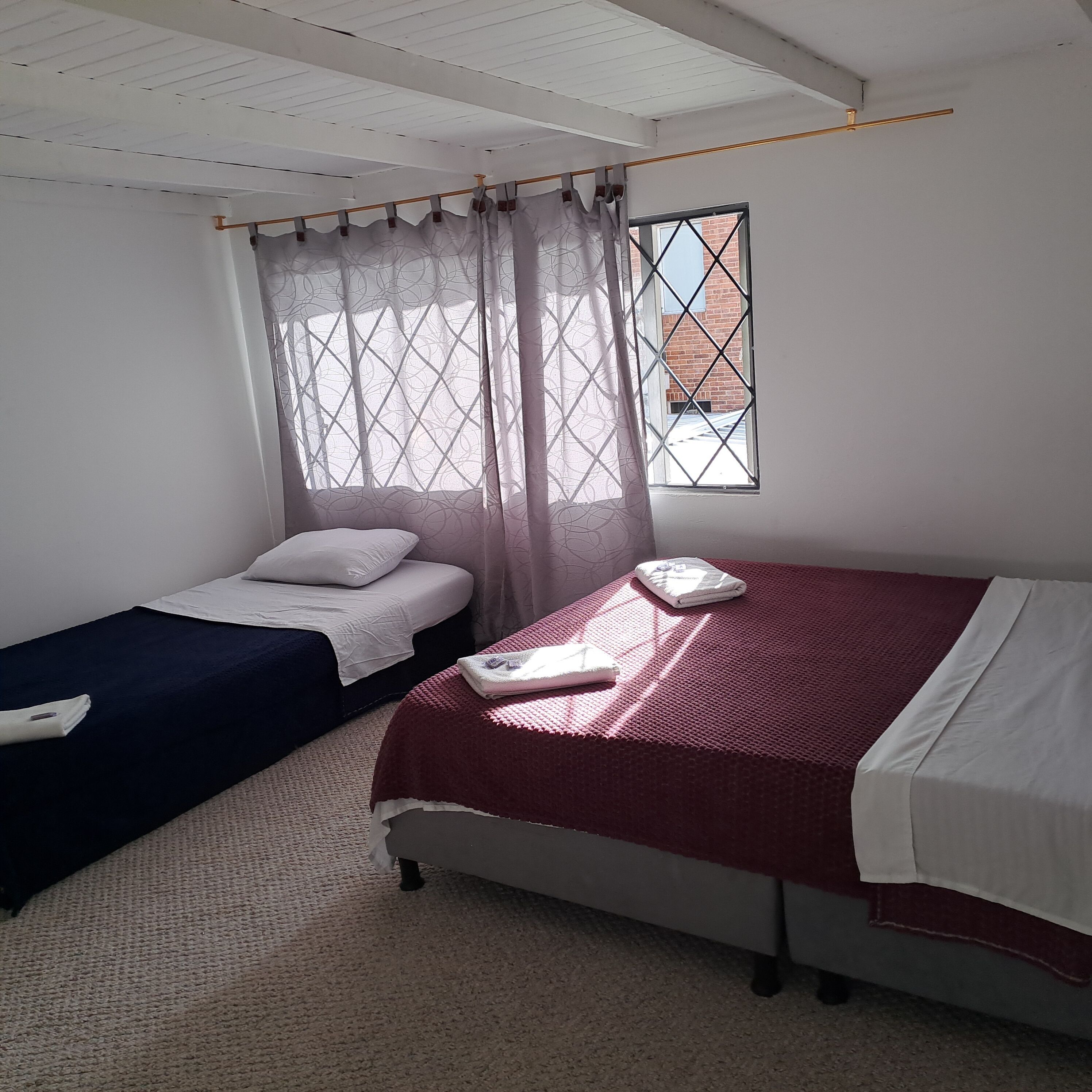 A room with two beds, one with a red bedspread and the other with a dark blue bedspread. There is a window with a view of a brick building outside.