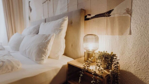 A neatly made bed with white linens and pillows, a bedside table with a lamp and decorative plants, and a wall-mounted shelf with a rustic design. - Staylight | City Loft in the Old Town | Balcony (Hildesheim)