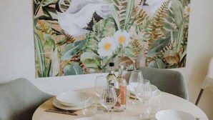 A dining table set with plates, glasses, and cutlery, surrounded by grey chairs, against a backdrop of a tropical-themed painting. - Staylight | City Loft in the Old Town | Balcony (Hildesheim)