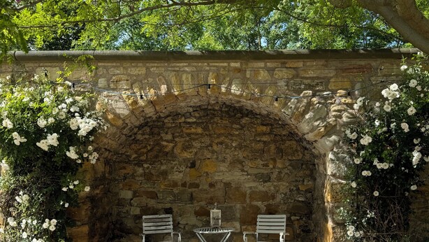 Un arco de piedra con dos sillas de metal y una pequeña mesa, rodeado de flores blancas y follaje.
