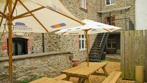 A stone building with a wooden staircase, outdoor seating with picnic tables, and umbrellas with logos. - The Red Lion (Knighton)