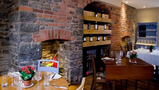 A restaurant with a stone wall and brick fireplace, wooden shelves with wine bottles, and a table set for dining. - The Red Lion (Knighton)