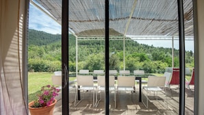 Un patio techado con una mesa y sillas, una planta en maceta y vista a una ladera boscosa.