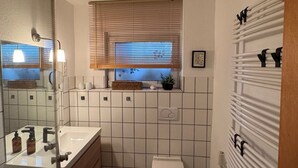 A compact bathroom with a white sink, mirror, and toilet. There is a window with blinds, a ceiling fan, and a radiator. - Spacious vacation apartment in Leverkusen-Opladen, quiet but centrally located (Leverkusen)
