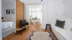 Een moderne hotelkamer met een houten scheidingswand, een witte bank, een televisie, een zithoek met een tafel en stoelen, en een witte deur.