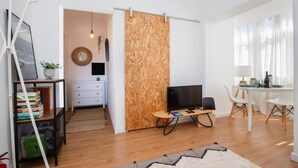 Een moderne woonkamer met een televisie, een wand van houten panelen, witte meubels en een eethoek met een tafel en stoelen.
