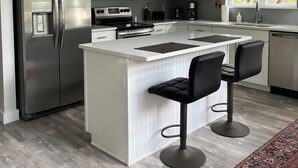 Une cuisine moderne avec des meubles blancs, des appareils en acier inoxydable, un îlot central doté de tabourets et une grande fenêtre avec des stores.