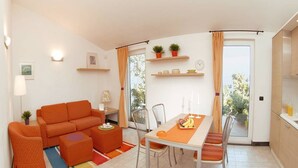 Een moderne woonkamer met een bank, een voetenbankje en een eettafel met stoelen. Er staan planten in potten op planken, een klok en een raam met gordijnen.