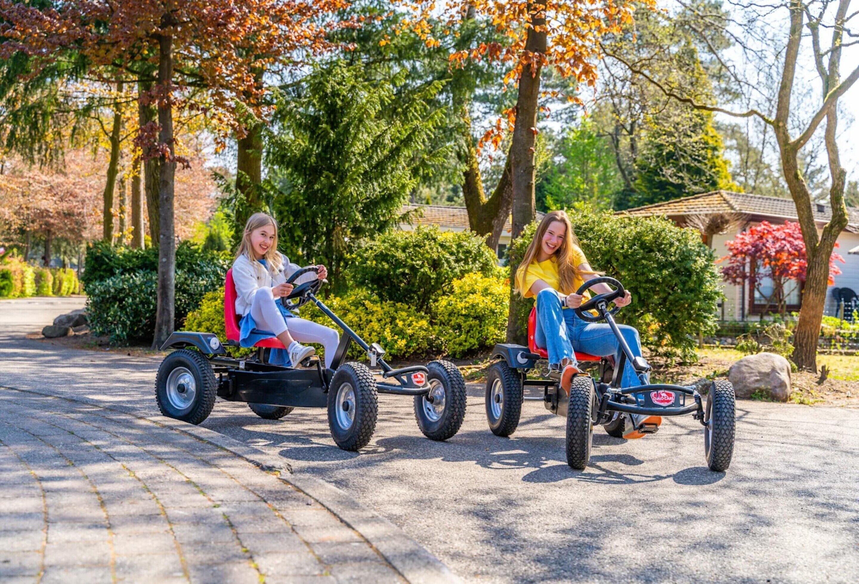 Dos personas manejando karts sobre un camino pavimentado en un parque con árboles y un edificio al fondo.