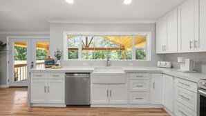 A modern kitchen with white cabinetry, a stainless steel dishwasher, a large window with a view of greenery, and a wooden deck outside. - Lakeview Retreat | Family-Friendly · Near Downtown (Macon)