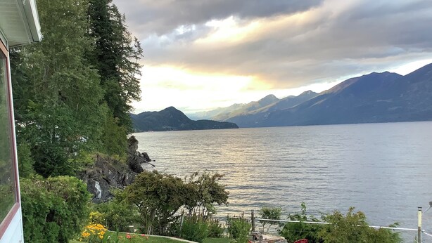 A lakeside view with mountains in the background, a grassy area with a pathway, and a chair. - Vacation Home For Rent - Ainsworth BC (Ainsworth)